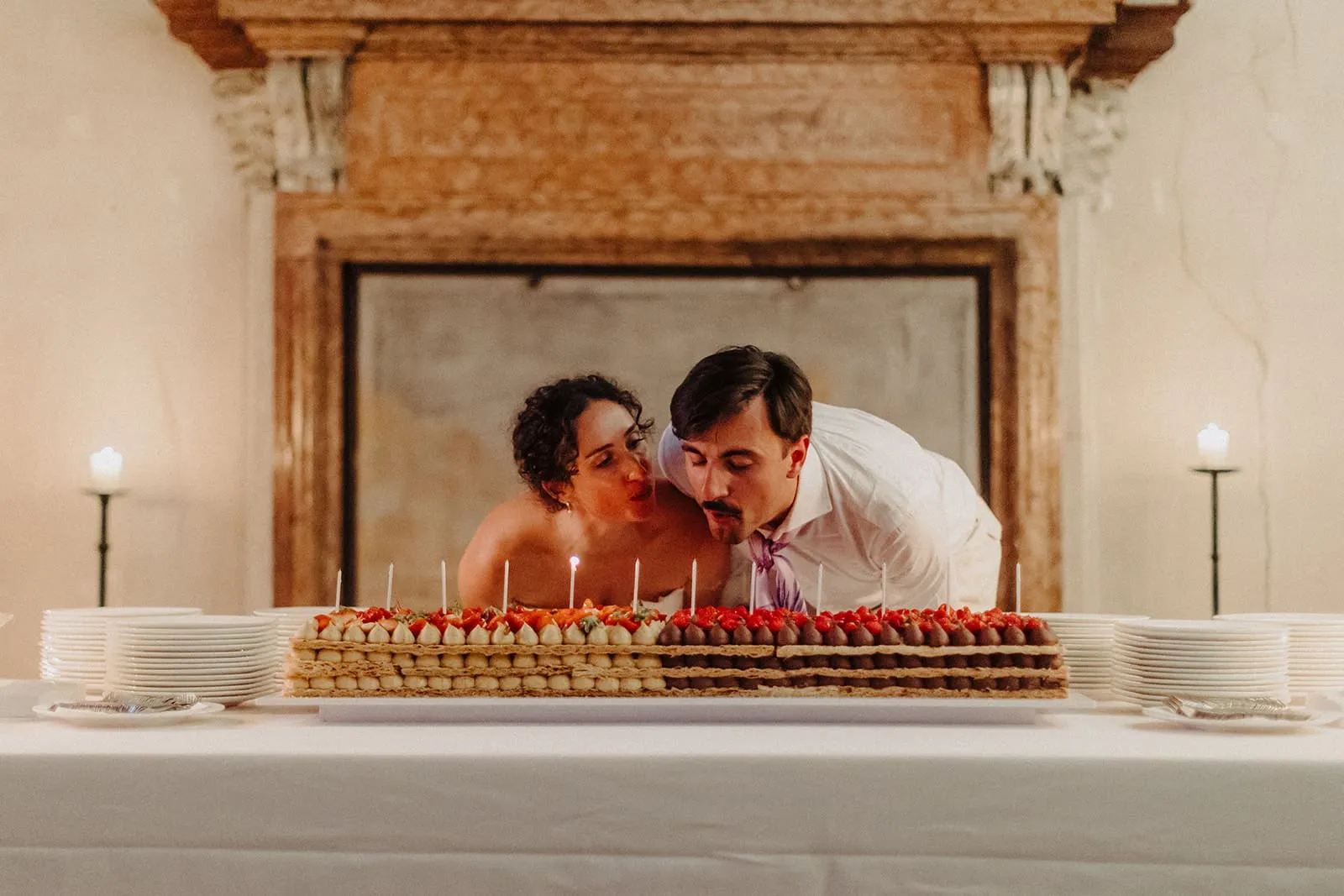 The couple leaning over the candlelit cake in front of the stone fireplace