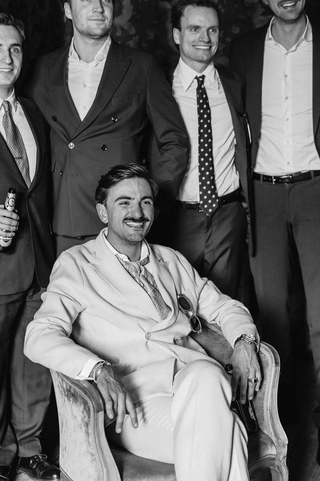 The groom sitting in the armchair, his friends standing behind him