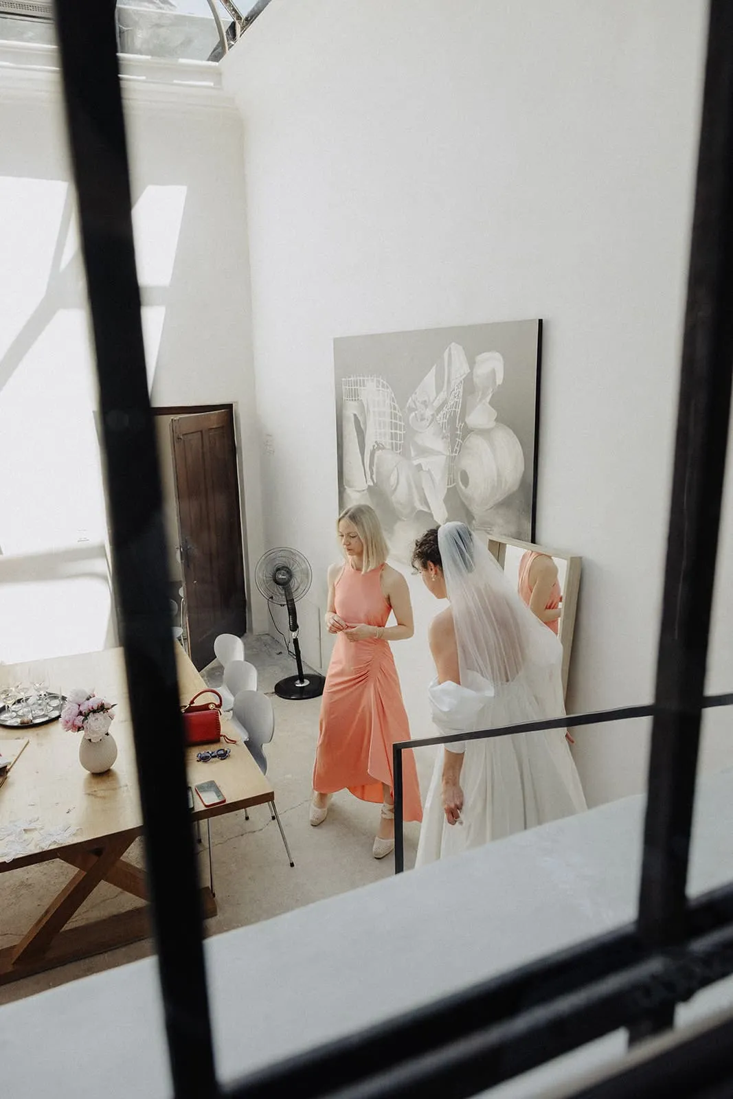 The bride in her veil through a window, a friend in orange beside her