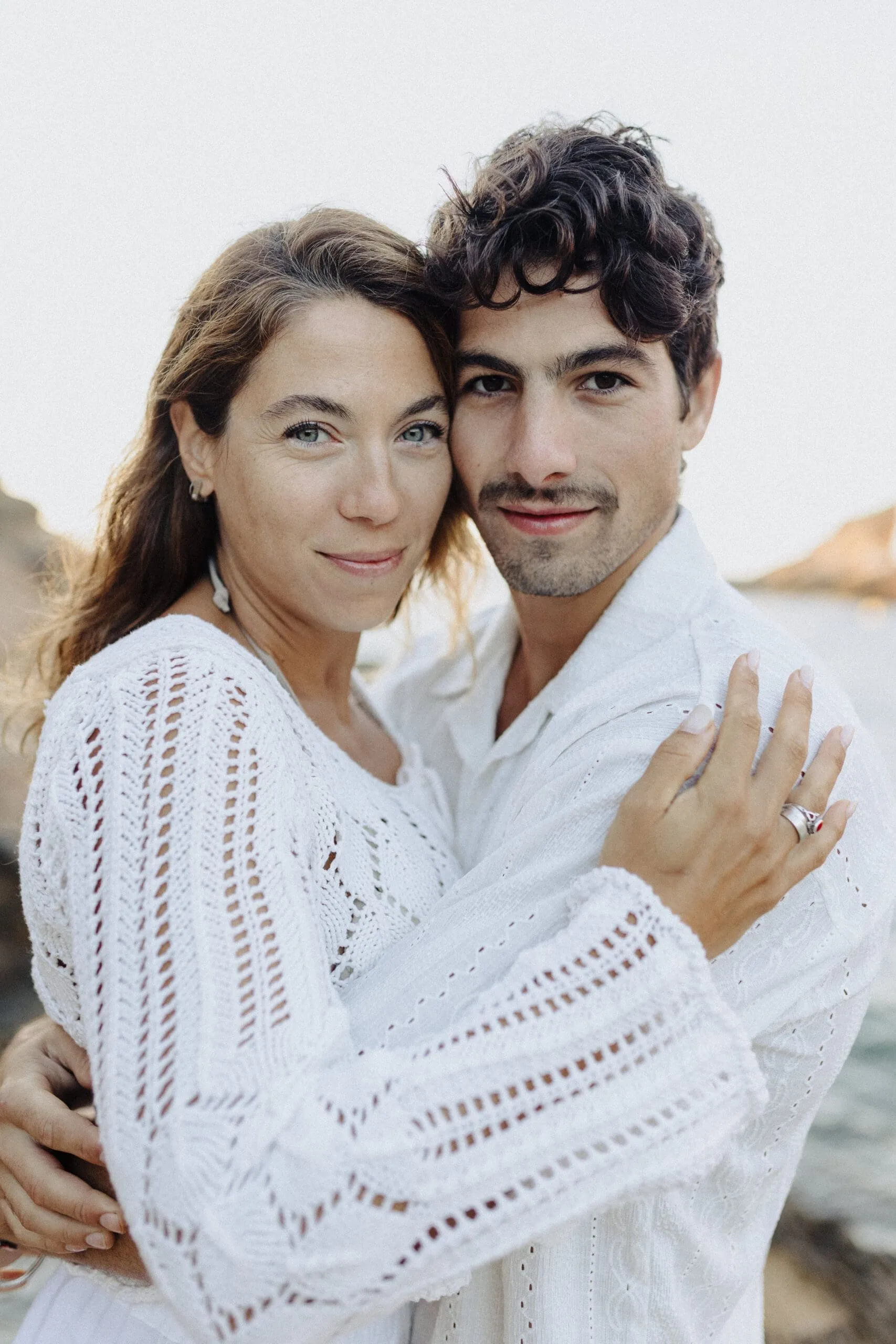 A couple in white knitwear holding each other close by the sea