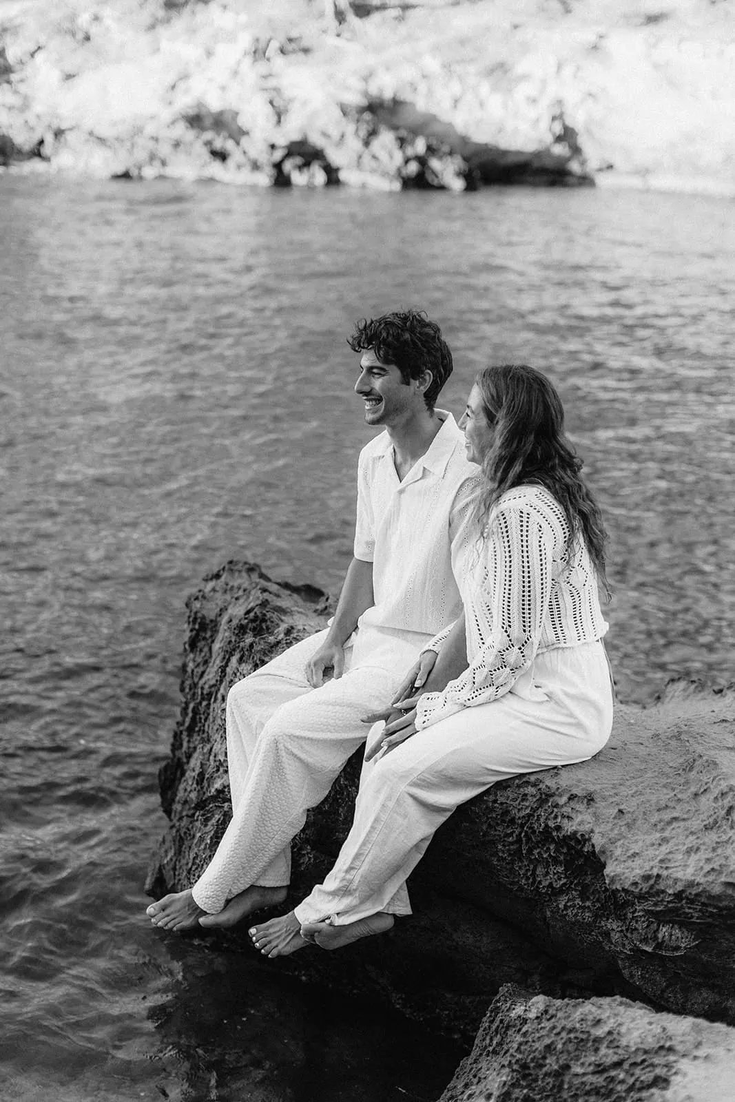 The couple sitting barefoot on a rock above the water, laughing