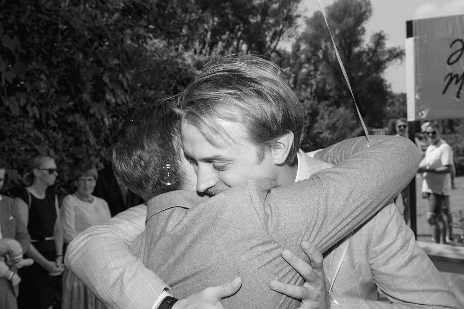 The groom holding a friend in a long hug outside, guests watching