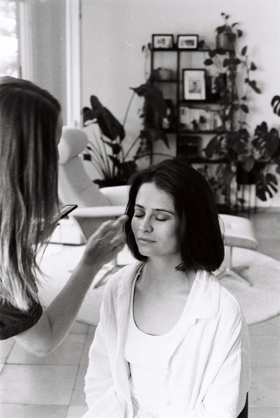 A brush at the bride's temple, her eyes closed, plants on the shelf behind
