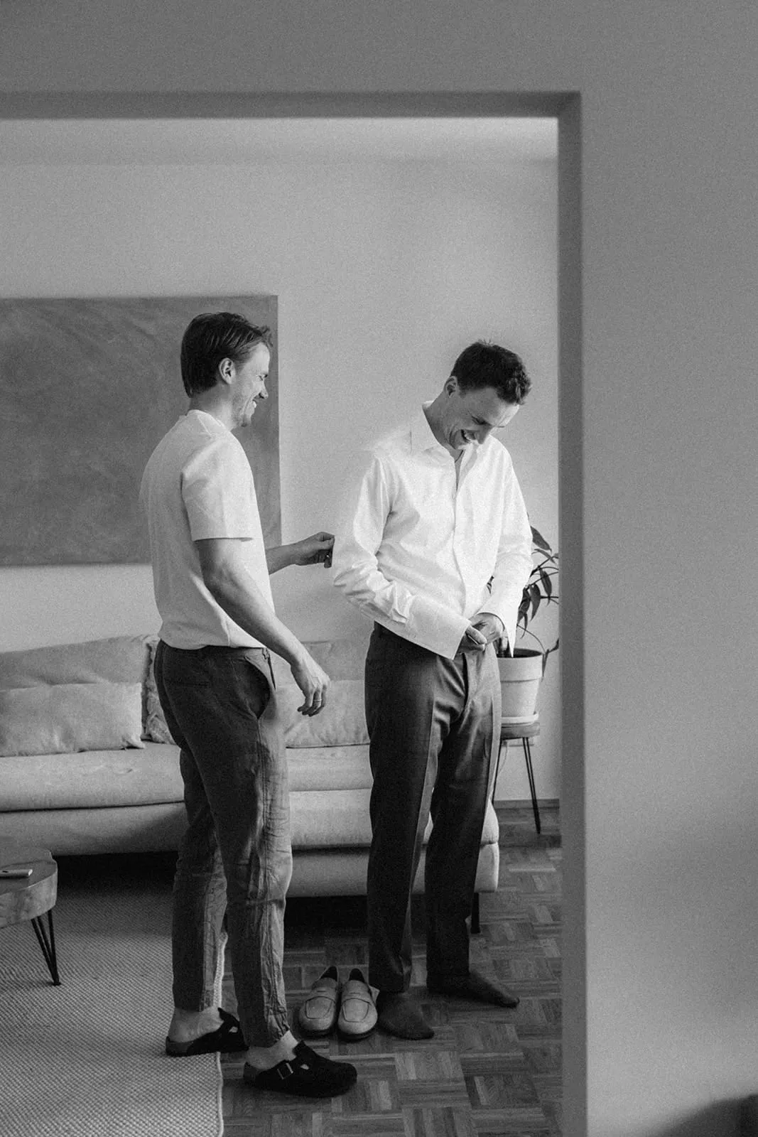 A friend helping the groom with his shirt in a bright living room
