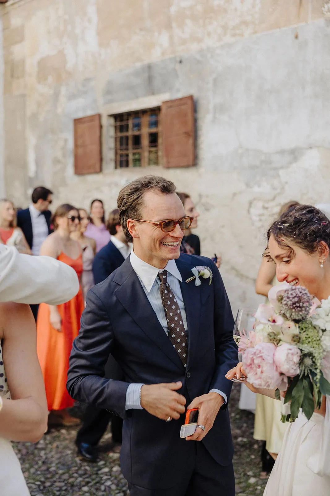 A guest laughing in the courtyard, the bride with her bouquet at the edge